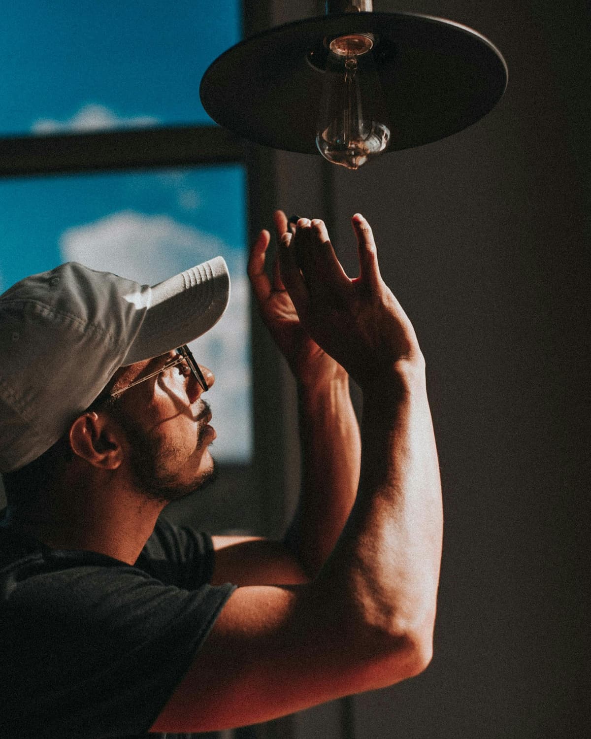 Man in a cap reaching up to install a pendant light fixture near a bright window